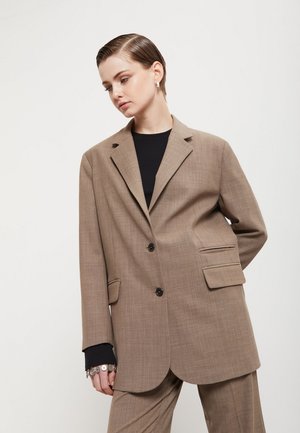 Donna con capelli corti che indossa un blazer marrone oversize e pantaloni abbinati sopra una maglia nera, guarda verso il basso con una mano dietro la schiena.
