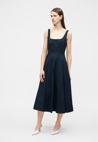 Γυναίκα με ίσια καστανά μαλλιά που φοράει μια αμάνικη μεσάτη μπλε ροζ φόρεμα με τετράγωνη λαιμόκοψη και λευκές μυτερές γόβες, στέκεται σε λευκό φόντο.