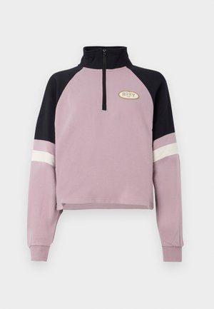Pull de coupe crop en violet avec un col noir et des accents de manches. Présente une fermeture éclair à mi-hauteur et le logo Roxy en doré sur la poitrine. Texture douce.