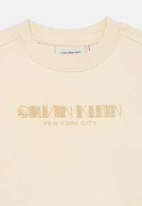 Felpa color crema con logo testurizzato in glitter dorato "Calvin Klein New York City". Presenta un colletto rotondo e maniche a costine.