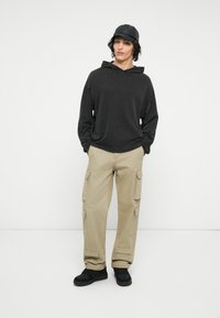 Sort hættebluse, beige cargo-bukser med lommer på siderne og sorte sneakers. Modellen bærer en sort kasket og står foran en hvid baggrund.