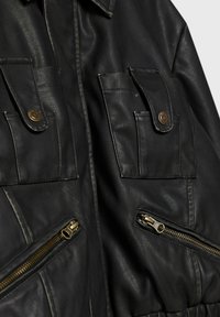 Černá kožená bunda se dvěma náprsními kapsami, zlatými kovovými doplňky a dvěma šikmými zipovými kapsami. Texturovaný povrch a viditelné švy.