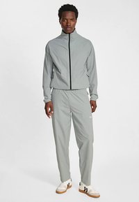 Lichtgrijs trainingspak met zwarte rits, zakken met rits en logodetail. Draagt witte sneakers met zwarte strepen.