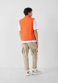 Oranje puffer vest met een doorgestikt ontwerp, gecombineerd met een wit shirt en beige cargobroek. De persoon draagt grijze sneakers.