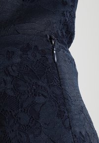 Robe marine avec une superposition en dentelle texturée et une fermeture éclair latérale. Le tissu présente une finition lisse avec des motifs en dentelle complexes.