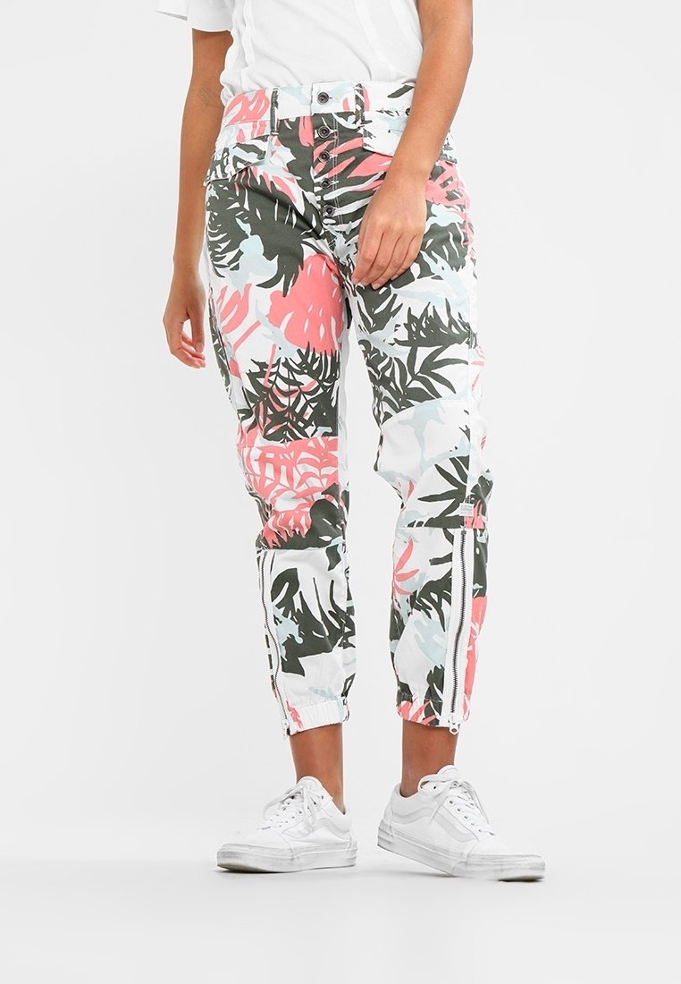 Mönstrade joggerbyxor med grön, rosa och vit tropisk lövdesign. Har elastiska muddar och dragkedjedetaljer. Bärs med vita sneakers.