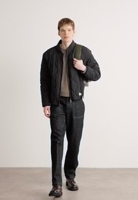 Crna bušena bomber jakna, sivi pulover, tamne hlače i sjajne cipele. Model nosi zeleni ruksak, stoji protiv svijetle pozadine.