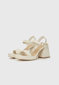 Sandales à talon bloc blanches avec deux larges brides et une sangle de cheville réglable dotée d'une boucle dorée. Texture lisse et design minimaliste.