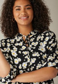 Blusa nera a fiori con margherite bianche e centri gialli, maniche corte e chiusura a fiocco sul collo. Tessuto morbido.