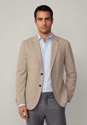 Beige Wollblazer mit zwei schwarzen Knöpfen, zwei frontalen Taschen und geschlitzten Revers, getragen über einem hellblauen Hemd und grauen schmal gestreifen Hosen.