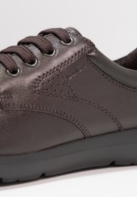 Close-up van een donkerbruin lederen sneaker met dikke bruin laces, gedetailleerde stiksels, geperforeerde accenten en het "GEOX"-logo op het zijpaneel.
