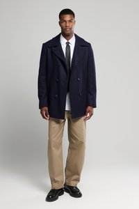 Abrigo marinero de lana en corte de doble botonadura, con botones grandes, llevado sobre una camisa blanca y una corbata negra, combinado con pantalones caqui.