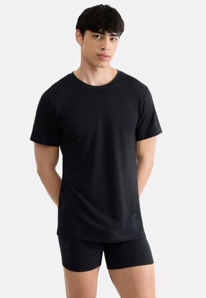 Młody mężczyzna stoi, ubrany w prostą czarną koszulkę z krótkim rękawem i dopasowane czarne spodenki, na neutralnym tle.