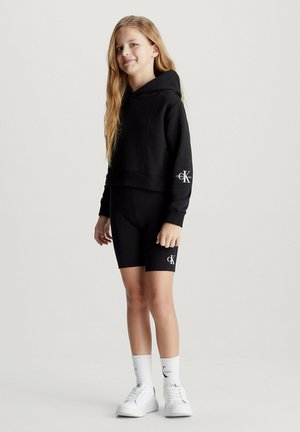 Cropped hoodie nera abbinata a pantaloni da bicicletta con logo, insieme a sneakers bianche e calze bianche a coste. Tessuto liscio.