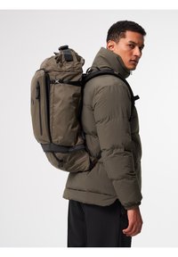 Khaki-Rucksack, der von einer Person getragen wird, aus glattem, strapazierfähigem Stoff gefertigt, mit mehreren Fächern und schwarzen Akzenten. Gepolsterte Schultergurte sind sichtbar.