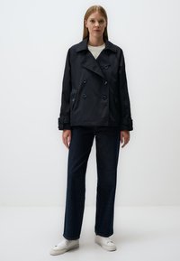 Granatowy, dwurzędowy trench z dużymi guzikami, bocznymi kieszeniami i szerokimi wyłogami, zestawiony z prostymi ciemnymi dżinsami i białymi butami.