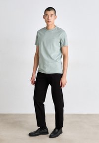 Világos zöld, rövid ujjú póló, kerek nyakkivágással, fekete nadrággal és fekete slip-on cipővel párosítva, viseli egy álló férfi modell.