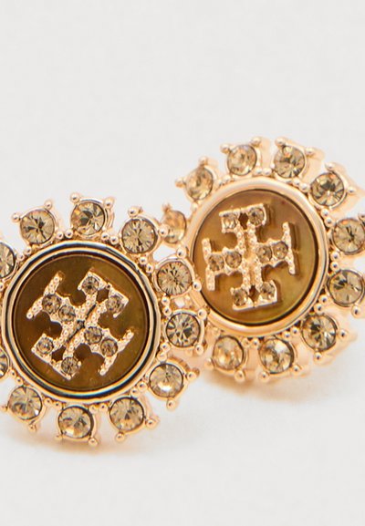 Brincos redondos dourados com cristais transparentes ao redor de um emblema central feito de pequenas pedras brilhantes dispostas em um padrão semelhante a uma cruz.