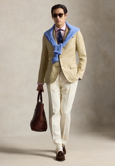 Homme portant un costume beige, un pull bleu drapé sur les épaules, des lunettes de soleil et portant un sac en cuir marron en marchant.