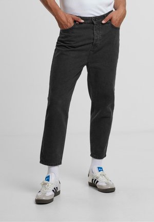 Person, die schwarze, schmal zulaufende Jeans trägt, mit Händen in den Taschen, weißen Socken und weißen Turnschuhen mit schwarzen Streifen und blauen Stern-Logos.