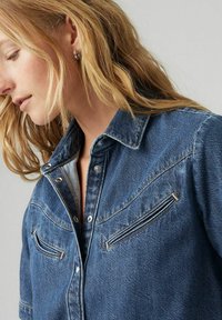 Camicia in denim blu scuro con chiusura a bottoni, due tasche sul petto con cerniere verticali e dettagli di cucitura a contrasto lungo le cuciture.