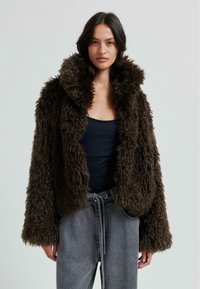 Brun texturerad fuskpälsjacka med hög krage och vida ärmar, kombinerad med mörkgrå dragsko-byxor, på en neutral bakgrund.