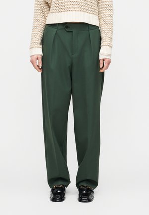 Persona che indossa pantaloni verde scuro a gamba larga, mocassini neri con fibbia dorata e un maglione beige lavorato a maglia con maniche risvoltate.