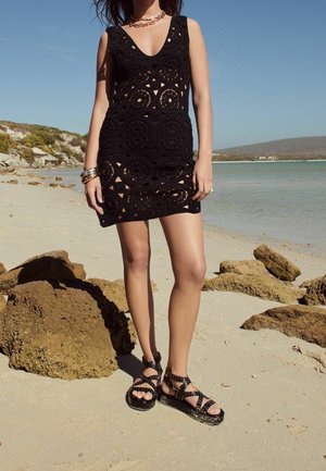 Femme debout sur une plage de sable portant une mini robe en crochet noire et des sandales noires cloutées près de grands rochers au bord d'une mer calme.