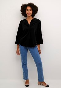 Chemisier noir à manches longues avec un décolleté en V et des détails de pli subtils, associé à un jean bleu clair et des chaussures beiges avec des accents noirs sur les orteils.
