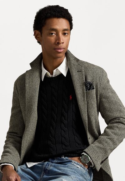Blazer gris à motif chevrons sur un pull en maille torsadée noir, chemise blanche à col et jeans bleu déchirés. Comprend un mouchoir de poche et une montre.