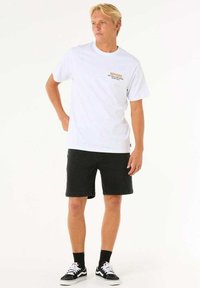 T-shirt bianca in cotone con testo dorato stampato, abbinata a pantaloni corti di jeans neri. Modello che indossa calze nere e sneaker bianche e nere.