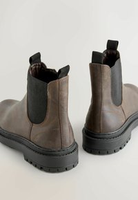 Rudos odinės kulkšnės batai su elastingais šoniais, apvaliu pirštu ir juodu guminiu padu, turinčiu tekstūruotą bėgimo raštą. Matomas siūlų siuvimo detalė.