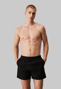 Costumi da bagno neri realizzati in tessuto leggero. Presentano un elastico in vita con cordoncino e tasche laterali. Design semplice senza pattern.