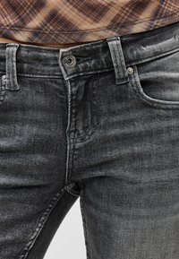 Κοντινό πλάνο ξεθωριασμένου τζιν μαύρου χρώματος με κουμπί και φερμουάρ, φορεμένα με καφέ καρό πουκάμισο που είναι φουσκωμένο μέσα.