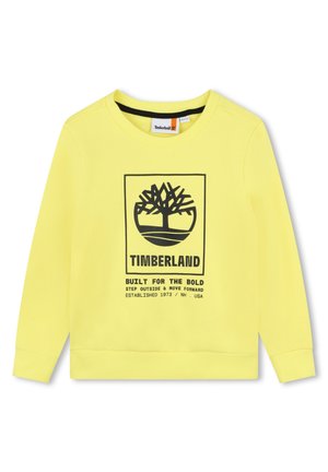 Sweatshirt jaune en tissu doux avec un graphique noir représentant un arbre et le texte : "TIMBERLAND CONÇU POUR LES AUDACIEUX," avec des poignets côtelés.