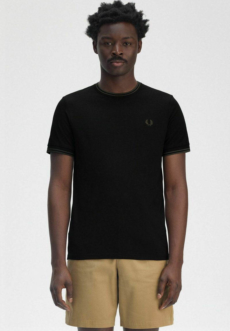 Camiseta negra con cuello redondo, ribete verde y pequeño logo bordado en el pecho, combinada con pantalones cortos beige.