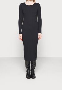 Vestido negro de manga larga con cuello redondo, silueta ajustada y largo hasta la rodilla, combinado con botas altas negras.