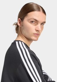 Μαύρο φούτερ με λευκό σχέδιο τριών ρίγων στα μανίκια, με στρογγυλή λαιμόκοψη και υφασμάτινη υφή. Το μοντέλο έχει μεσαίου μήκους μαλλιά και σκουλαρίκι.