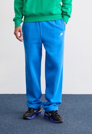 Man draagt felblauwe Nike joggingbroek, groene hoodie en zwarte Nike sneakers, staand op een blauw tapijt.