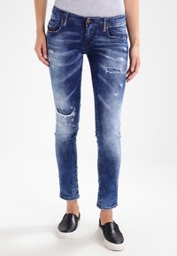 Modré džíny skinny s vydřeným vzhledem, s roztrženými vzory, pěti kapsami a středním pasem. Nosené s černými slip-on botami.