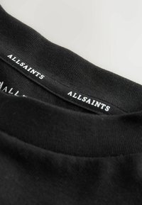 Schwarzer Baumwoll-T-Shirt-Kragen mit glatter Textur. Auf der Innenseite des Halsausschnitts befindet sich ein weiß aufgedrucktes "ALLSAINTS"-Logo. Schlichtes Design.
