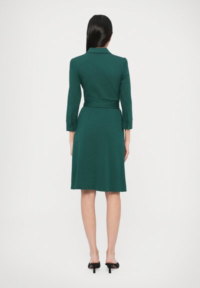 Πράσινο φόρεμα μέχρι το γόνατο με εφαρμοστό μπούστο και φούστα που φαρδαίνει, μακριά μανίκια, μανσέτες με κουμπιά και ζώνη στη μέση. Λείο, απαλό ύφασμα.