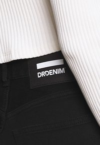 Os jeans de ganga pretos têm um rótulo tecido com "DRDENIM" em branco, parcialmente coberto por uma parte de cima branca canelada. O tecido parece liso.