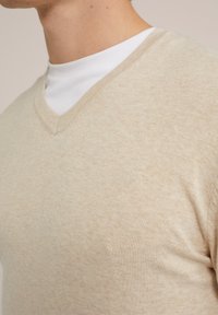 Beige stickad tröja med V-ringning, som har en mjuk textur och något melerad yta. Lager på en vit tröja med rund hals.