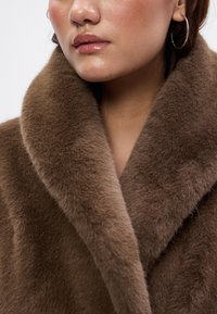 Casaco de pelo castanho com um colarinho largo e peludo e uma textura suave, apresentando uma vista aproximada do brilho natural e um leve reflexo.