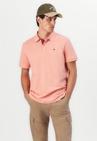 Polohemd aus hellrosa Baumwolle mit einer Drei-Knopf-Leiste, kurzen Ärmeln und einem kleinen Logo auf der Brust. Khaki Cargo-Hose.