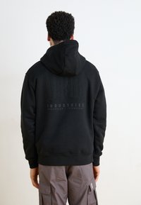 Sort hættetrøje med forhøjet skrift på ryggen: "ALPHA INDUSTRIES" og "KNOXVILLE TENNESSEE." Stoffet ser blødt og holdbart ud.
