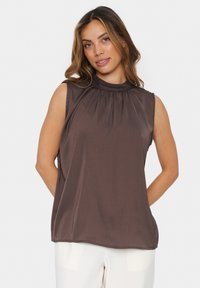 Top senza maniche di colore marrone con collo alto e dettagli arricciati, caratterizzato da una superficie liscia e una vestibilità rilassata, abbinato a pantaloni color crema.