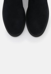 Schwarze Wildleder-Stiefeletten mit runder Zehenpartie, glatter Textur und strapazierfähiger Gummisohle, von oben gegen einen neutralen Hintergrund betrachtet.