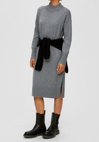 Abito grigio in maglia con scollo a coste e spacchi laterali, abbinato a un maglione nero legato in vita e stivaletti neri alla caviglia.
